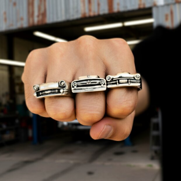 Drei massiv gearbeitete Silberringe im Design klassischer US-Car Frontgrills auf einer geballten Faust vor Werkstatt-Hintergrund – handgefertigter Biker- und Auto-Schmuck für Gearheads