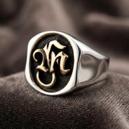 Eleganter Silberring mit goldfarbenem Monogramm, dunkler strukturierter Siegelplatte und polierter Ringschiene auf weichem Stoff