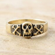Goldener Skull Ring mit plastischem Totenkopf, gekreuzten Knochen und geschwärzten Details auf hellem Holz