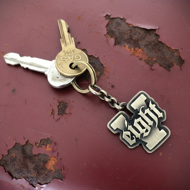 V8 Schlüsselanhänger Oldschool – Messing, „V-eight“ Frakturlogo Oldschool V8-Schlüsselanhänger aus Messing mit „eight“-Frakturlogo am Schlüsselbund, fotografiert auf bordeauxroter, rostpatinierter Motorhaube.