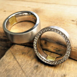 Handgefertigte Trauringe aus 585 Weißgold auf Holz, einer mit Brillant-Pavé-Fassung, beide mit sichtbarer Gelbgold-Einlage innen für Fingerabdruck